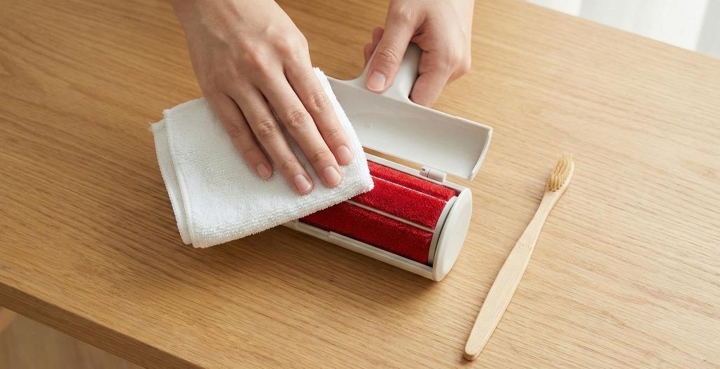 An overhead photograph showing a pair of hands carefully cleaning the red velvet roller surface of an open white ChomChom pet hair remover using a white cotton cloth. A bamboo toothbrush rests beside the roller on a wooden table surface.